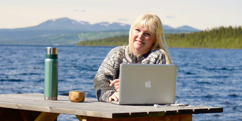 Pernilla Öberg vid ett träbord utomhus framför havet med sin Mac-dator