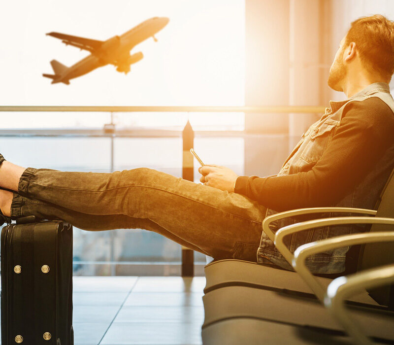 En kille sitter på en stol på flygplatsen med sin mobil och tittar drömskt ut mot ett lyftande plan.